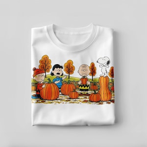 Pode incluir: T-shirt branco de gola redonda com uma ilustração colorida dos personagens de Peanuts em um campo de abóboras. Charlie Brown, Lucy, Linus e Snoopy são mostrados com abóboras e folhas de outono. Um design festivo para o outono.