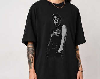 May include: A black short-sleeved t-shirt featuring a high-contrast, monochromatic graphic print of a person wearing sunglasses and a sleeveless top. The graphic is positioned centrally on the chest, showcasing a detailed portrait style illustration.