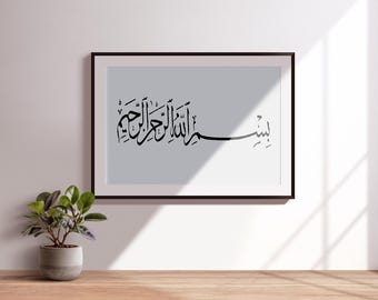 May include: Framed art print featuring black Arabic calligraphy on a light gray background. The calligraphy is set within a black frame, creating a striking contrast. A small potted plant sits on a wooden surface in the foreground.