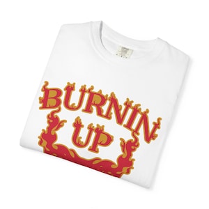 Könnte beinhalten: Gefaltetes weißes T-Shirt mit dem Schriftzug "BURNIN' UP" in roter und goldener Flammenoptik. Das Design zeigt eine feurige Grafik, die auf ein Thema von Hitze oder Energie hindeutet. Ein Etikett ist am Kragen sichtbar.