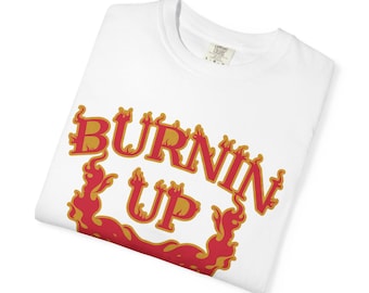 May include: White t-shirt with the words "BURNIN' UP" in red and gold flame-style lettering. The shirt is folded and has a tag on the collar. The design features a fiery graphic, suggesting a theme of heat or energy.