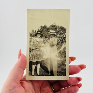 May include: Vintage black and white photograph of a couple in formal attire, likely from the early 20th century. The woman wears a patterned dress and hat, while the man is in a suit and hat. They are standing outdoors, possibly in a garden.