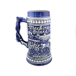 May include: A blue and white ceramic beer stein featuring a relief design of a horse-drawn carriage. The text Mackinac Island is embossed near the top, and the word MICHIGAN is embossed near the base. The handle has a textured, branch-like design.