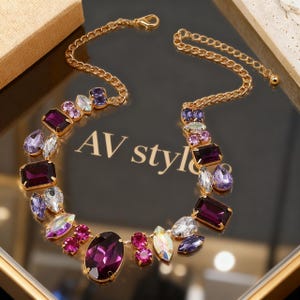 Puede incluir: Un collar de cadena en tono dorado con una variedad de cristales facetados en tonos morado intenso, lavanda claro y blanco iridiscente transparente. El collar incluye una piedra central ovalada de color morado y un cierre de mosquetón con el texto AV style.