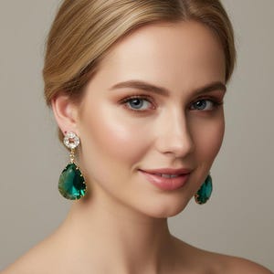 Può includere: Primo piano di una donna con orecchini dorati. Ogni orecchino presenta una parte superiore quadrata trasparente e una grande gemma verde a forma di goccia. Gli orecchini sono un accessorio glamour. Capelli biondi, occhi blu.