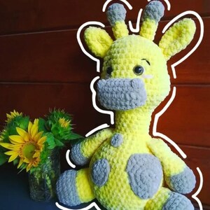 May include: A handmade yellow and gray crochet giraffe plush toy with gray spots and accents. The giraffe has large eyes and is sitting. A vase of sunflowers is to the left of the giraffe.