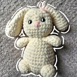 May include: A handmade, cream-colored crochet bunny with black eyes and a pink nose. The bunny has long, floppy ears and a small pink bow. The plush toy is soft and cuddly, perfect for a child's room or as a decorative item.
