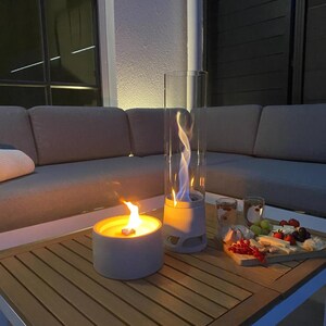 May include: Two outdoor tabletop fire pits with flames, one round and one tall, cylindrical, on a wooden table. A charcuterie board with fruit and cheese and two glasses are also on the table. A gray sofa is in the background.
