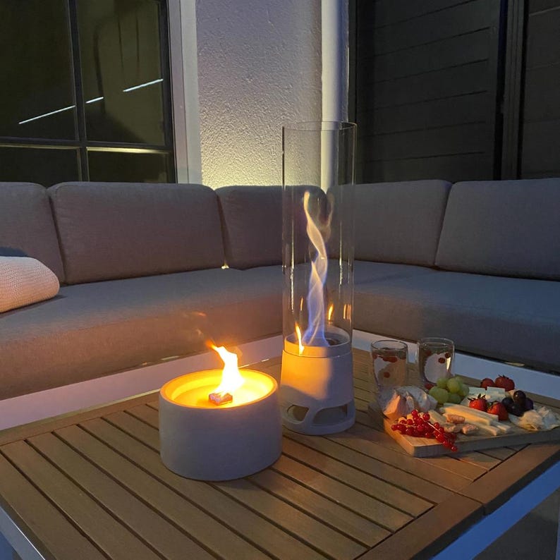 May include: Two outdoor tabletop fire pits with flames, one round and one tall, cylindrical, on a wooden table. A charcuterie board with fruit and cheese and two glasses are also on the table. A gray sofa is in the background.