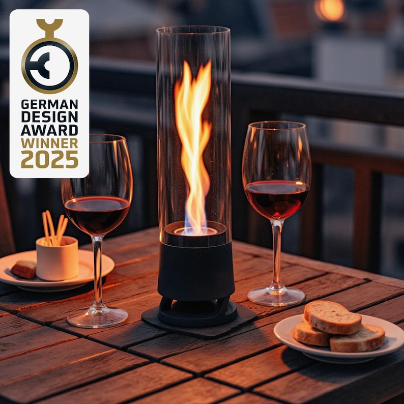 May include: A black and clear glass tabletop fireplace with a flame, flanked by two wine glasses filled with red wine. The table also holds a small plate of bread and a small dish with snacks. The image includes a "German Design Award Winner 2025" logo.