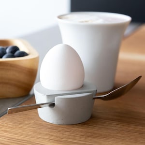 Può includere: Un uovo bianco in un portauovo di cemento grigio con un cucchiaio d'argento. Una ciotola di legno con mirtilli e una tazza di caffè bianca sono sullo sfondo. Gli oggetti sono su una superficie di legno.