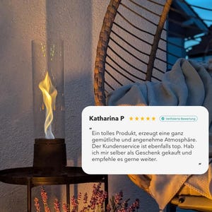 May include: A black outdoor tabletop fireplace with a glass wind guard, showcasing a dancing flame. The fire pit is set on a metal table with a lower shelf, and surrounded by purple flowers. A wicker chair is in the background.