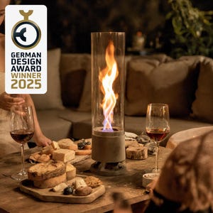 May include: A modern outdoor tabletop fireplace with a tall glass cylinder, showcasing a vibrant flame. The base is a neutral gray, and the scene includes wine glasses, a charcuterie board, and the text "GERMAN DESIGN AWARD WINNER 2025."