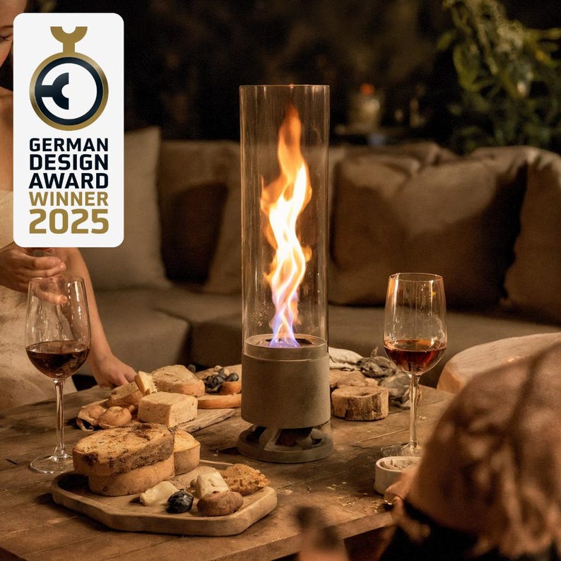 May include: A modern outdoor tabletop fireplace with a tall glass cylinder, showcasing a vibrant flame. The base is a neutral gray, and the scene includes wine glasses, a charcuterie board, and the text "GERMAN DESIGN AWARD WINNER 2025."