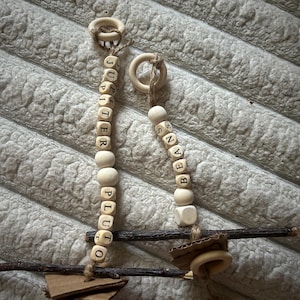 Puede incluir: Dos juguetes colgantes de cuentas de madera. Uno deletrea "Jupiter" y el otro "Beans". Cada uno tiene un anillo de madera en la parte superior y está unido a una pequeña base de cartón y madera. Las cuentas son de color madera natural.