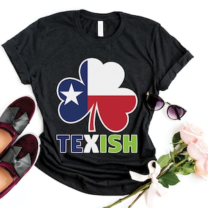 Könnte beinhalten: Dunkelgraues T-Shirt mit einem Kleeblatt-Design in den Farben der Texas-Flagge. Das Wort "TEXISH" ist darunter in Blau und Grün gedruckt. Das Bild enthält auch bordeauxfarbene Loafer, Sonnenbrillen und Rosen.