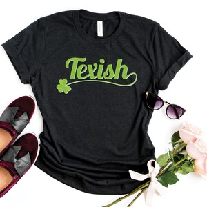 Könnte beinhalten: Dunkelgraues T-Shirt mit dem Wort "Texish" in grüner Schrift und einem grünen Kleeblatt. Das Shirt ist mit bordeauxfarbenen Samt-Loafers mit schwarzen Schleifen, Sonnenbrille und rosa Rosen auf weißem Hintergrund gestylt.