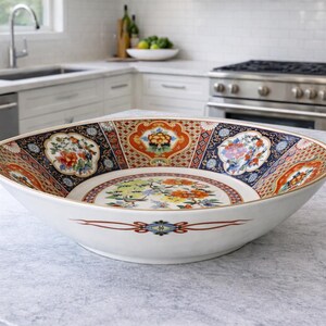 Peut inclure: Grand saladier ovale en céramique blanche, orné de motifs floraux et géométriques complexes rouges, bleus, oranges et dorés. Le bol a un bord doré et est posé sur un plan de travail gris clair.