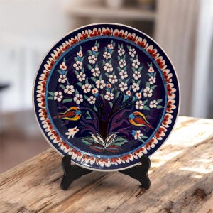 Peut inclure: Assiette décorative en céramique avec un fond bleu foncé et un motif d'arbre de vie. L'arbre présente des fleurs blanches et deux oiseaux colorés. Le bord a un motif rouge, bleu et blanc. L'assiette est présentée sur un support noir.