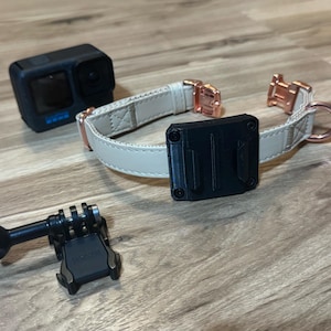 Op de afbeelding: Een zwarte actiecamera, een beige hondenhalsband met ros&eacute;gouden hardware en een zwarte camerabevestiging liggen op een houten oppervlak. De halsband heeft een zwarte montageplaat.