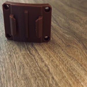 Op de afbeelding: Een bruine plastic houder met twee verticale lipjes en vier schroefgaten. De houder is rechthoekig met een gebogen bovenkant en is ontworpen om aan een oppervlak te bevestigen. Het item wordt getoond op een houten oppervlak.