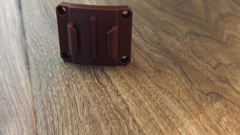 Op de afbeelding: Een bruine plastic houder met twee verticale lipjes en vier schroefgaten. De houder is rechthoekig met een gebogen bovenkant en is ontworpen om aan een oppervlak te bevestigen. Het item wordt getoond op een houten oppervlak.