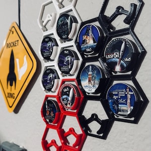 Puede incluir: Una colección de parches de misiones de cohetes ULA exhibidos en un soporte en forma de panal. Los soportes son blancos, negros y rojos. También es visible un letrero amarillo en forma de diamante con un gráfico de cohete.
