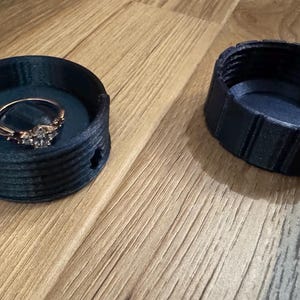 以下が含まれることがあります： ダークブルーの3Dプリントリングボックスと、お揃いの蓋。ボックスには、装飾的なデザインのゴールドリングが入っています。リングボックスは木製の表面に置かれています。