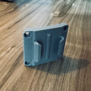 Op de afbeelding: Een grijze, rechthoekige 3D-geprinte houder met drie verhoogde secties en vier cirkelvormige gaten. De houder is ontworpen om een item vast te houden en ligt op een houten oppervlak.