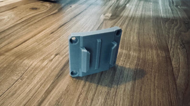 Op de afbeelding: Een grijze, rechthoekige 3D-geprinte houder met drie verhoogde secties en vier cirkelvormige gaten. De houder is ontworpen om een item vast te houden en ligt op een houten oppervlak.