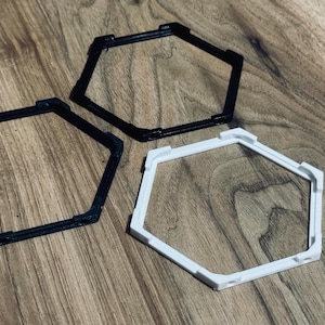 May include: Three geometric, hexagonal frames in black and white. The frames are made of a solid material and have a slightly raised edge. They are arranged on a wooden surface.