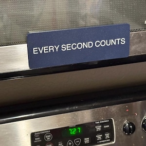 May include: A rectangular dark blue magnet with the white text EVERY SECOND COUNTS attached to the stainless steel door of a microwave oven, positioned directly above a stove control panel with a digital clock display showing 7:27.