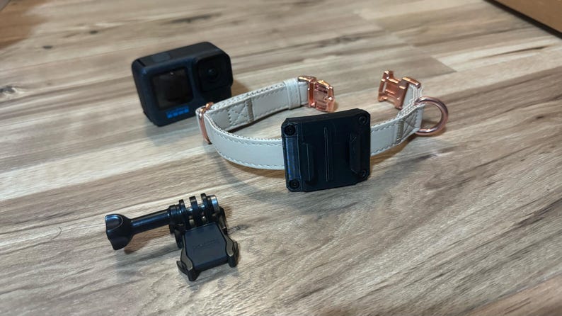 Op de afbeelding: Een zwarte actiecamera, een beige hondenhalsband met ros&eacute;gouden hardware en een zwarte camerabevestiging liggen op een houten oppervlak. De halsband heeft een zwarte montageplaat.