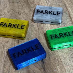 May include: Four colorful Farkle game boxes in yellow, gray, green, and blue are displayed on a wooden surface. Each box has the word "FARKLE" printed on the top. The boxes are rectangular and have a hinged lid.