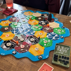 May include: A board game featuring a blue, irregular-shaped frame containing hexagonal tiles in yellow, green, brown, black, and white. Small circular number tokens are placed on the tiles. Two plastic card holders with text read ROAD DEV. CARD SETTLEMENT CITY.
