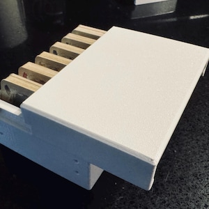 May include: A white rectangular box with a lid, containing several rectangular, light-colored objects. The box is on a dark surface. The objects inside the box appear to be stacked. The box has a textured surface.