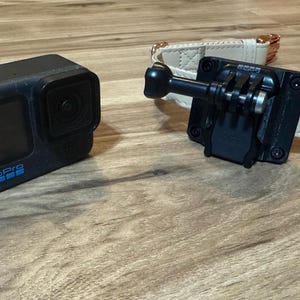 Op de afbeelding: Zwarte actiecamera met het woord "GoPro" in blauw. Een zwarte houder is bevestigd aan een beige hondenhalsband met ros&eacute;gouden hardware. De camera en de houder liggen op een houten oppervlak.