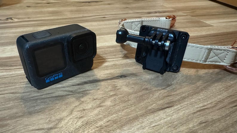 Op de afbeelding: Zwarte actiecamera met het woord "GoPro" in blauw. Een zwarte houder is bevestigd aan een beige hondenhalsband met ros&eacute;gouden hardware. De camera en de houder liggen op een houten oppervlak.