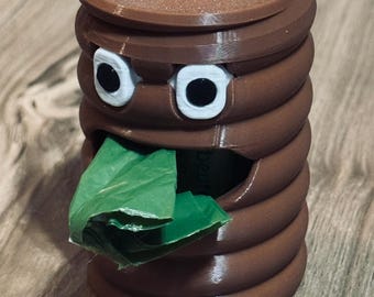 Op de afbeelding: Bruine, 3D-geprinte poepvormige dispenser met een deksel en een zilveren karabijnhaak. De dispenser heeft witte ogen met zwarte pupillen en een groen, bladachtig object dat uit de mond steekt. Op een houten oppervlak.