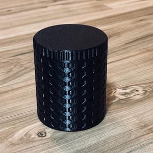 Puede incluir: Un objeto cilíndrico negro con una superficie texturizada y un patrón de panal. El objeto está sobre una superficie de madera. El objeto mide aproximadamente 10 cm de alto y 7,6 cm de diámetro.