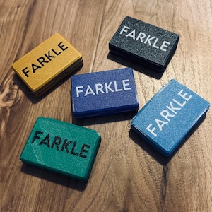 May include: Five rectangular game pieces with the word "FARKLE" in white text. The pieces are in various colors: yellow, black, blue, teal, and green. The pieces are arranged on a wooden surface.