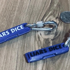 Può includere: Due portachiavi blu con la scritta "LIARS DICE" in lettere bianche. Ogni portachiavi ha un moschettone argentato. Una moneta è accanto ai portachiavi. I portachiavi sono su una superficie di legno.