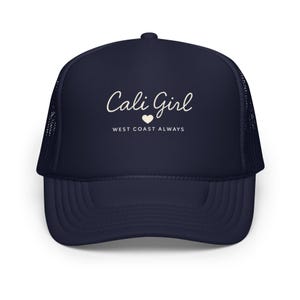 May include: Navy blue trucker cap with a curved peak and mesh back. The cap features the text "Cali Girl" in a cursive font, with a small heart and the words "West Coast Always" below.