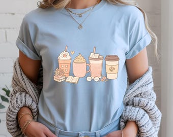 Puede incluir: Camiseta azul claro con una ilustración caprichosa de cuatro bebidas de café, libros y flores. Las bebidas están adornadas con crema batida, un corazón y una pajita. El diseño utiliza colores pastel suaves.