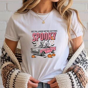 May include: White t-shirt with a Halloween-themed graphic. The design features a pink convertible car with skeleton passengers, pumpkins, bats, and the text "Get in, Loser we're getting Spooky."