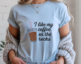 May include: Light blue t-shirt with the phrase "I like my coffee on the rocks" in black script. The graphic features a brown iced coffee cup with ice cubes and a pink straw. The shirt is a casual, everyday wear item.