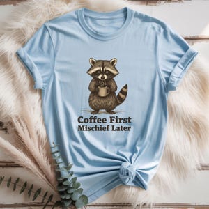 Op de afbeelding: Lichtblauw T-shirt met een cartoon wasbeer die een koffiekopje vasthoudt. De wasbeer is bruin en grijs met een gestreepte staart. De tekst "Coffee First Mischief Later" staat eronder.