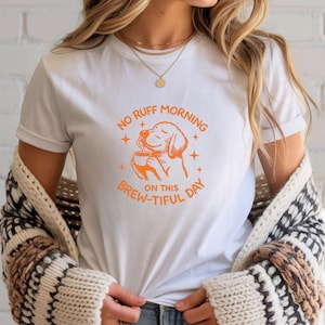 Op de afbeelding: Wit t-shirt met een oranje afbeelding van een hond die koffie drinkt. De tekst op het shirt luidt "NO RUFF MORNING ON THIS BREW-TIFUL DAY". Het shirt is gemaakt van een zacht materiaal en heeft korte mouwen.