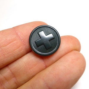 May include: A dark green plastic button with a raised white cross in the center. The button has a textured surface.