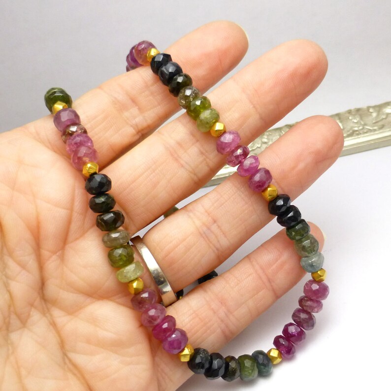 May include: A multi-colored gemstone necklace with pink, green, and black beads separated by gold spacers. The necklace is on a hand.
